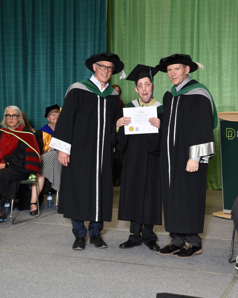 Brendan is in the middle, surrounded by two members of faculty. They are on stage at a convocation ceremony. Brendan is holding is certificate, wearing a grad cap and gown. The two faculty members on either side are wearing black ceremonial gowns and caps. 
